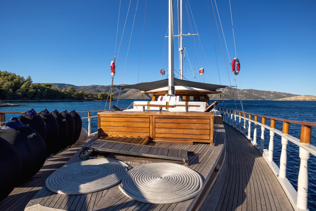 ABORDA View from the deck of a wooden yacht on calm blue water, with circular rugs, black fenders, lifebuoys, and distant green hills under a clear blue sky.