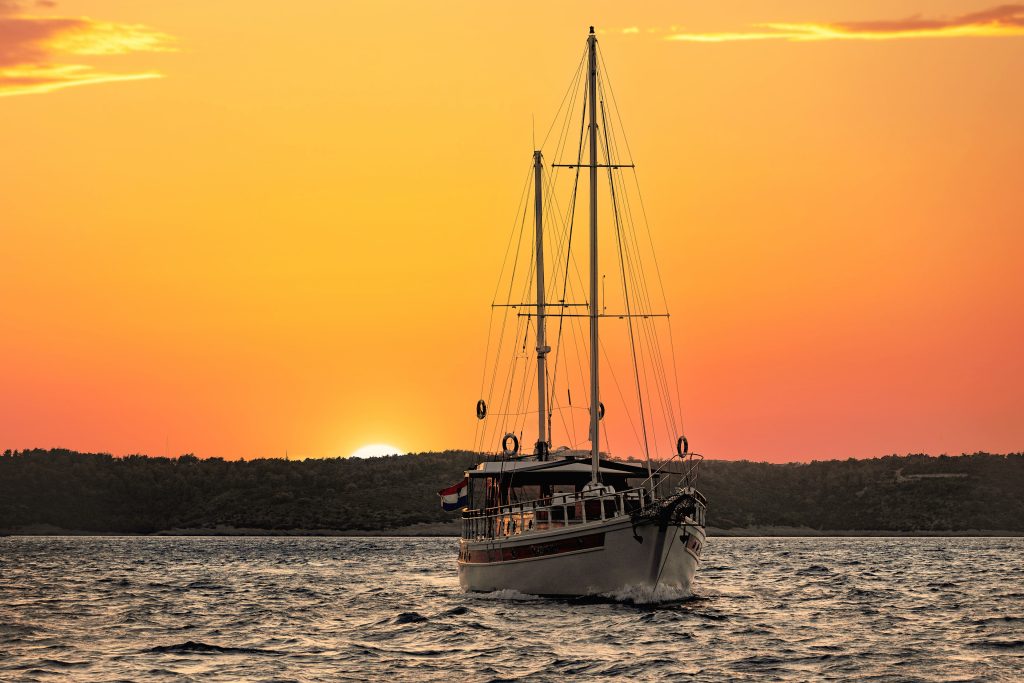 ABORDA A sailboat floats on calm water at sunset, with an orange sky and the sun setting behind a dark, tree-covered shoreline in the background.