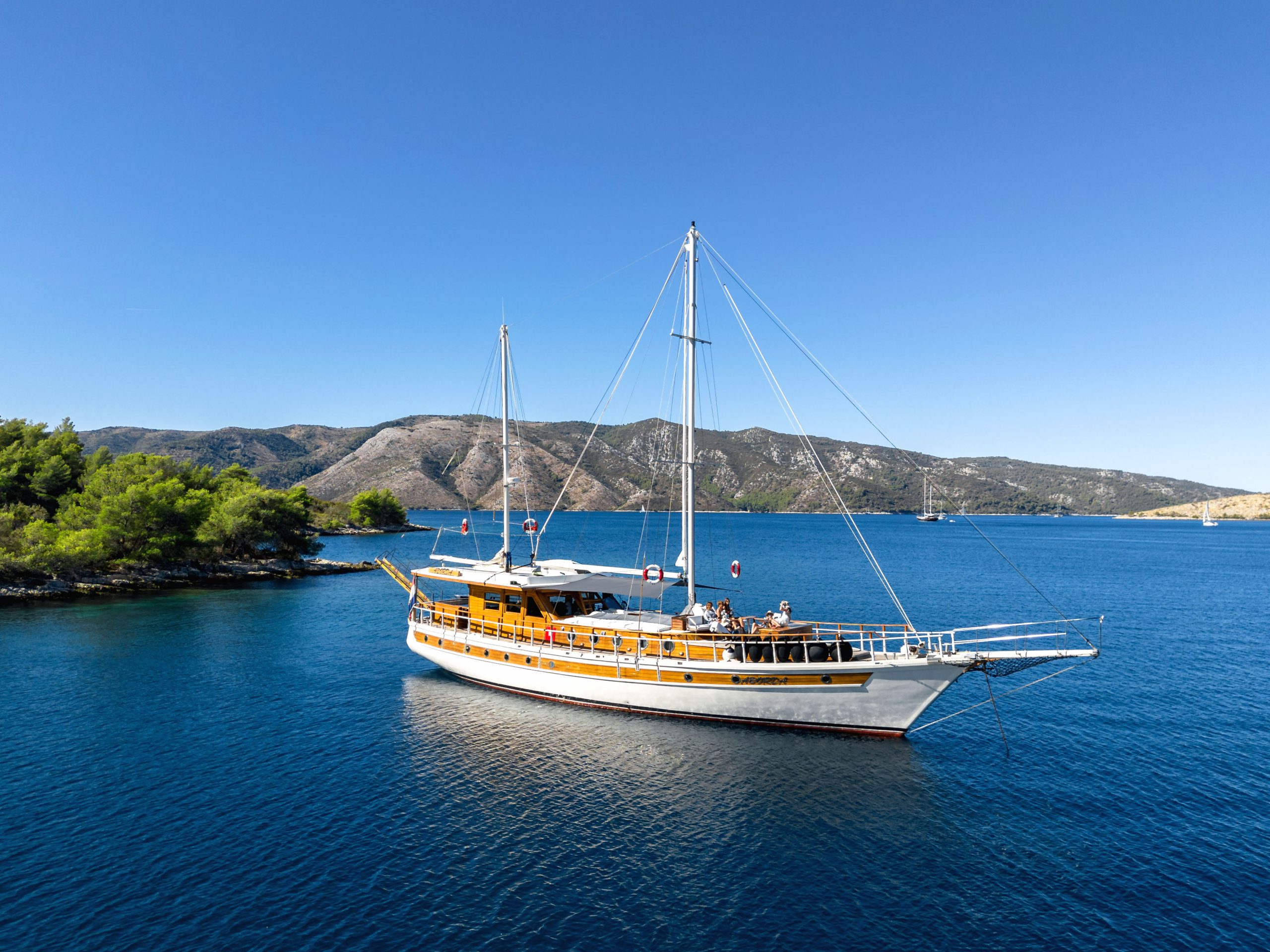 ABORDA A wooden sailboat with two masts floats on calm blue water near a tree-lined shore, with hills and a clear blue sky in the background.