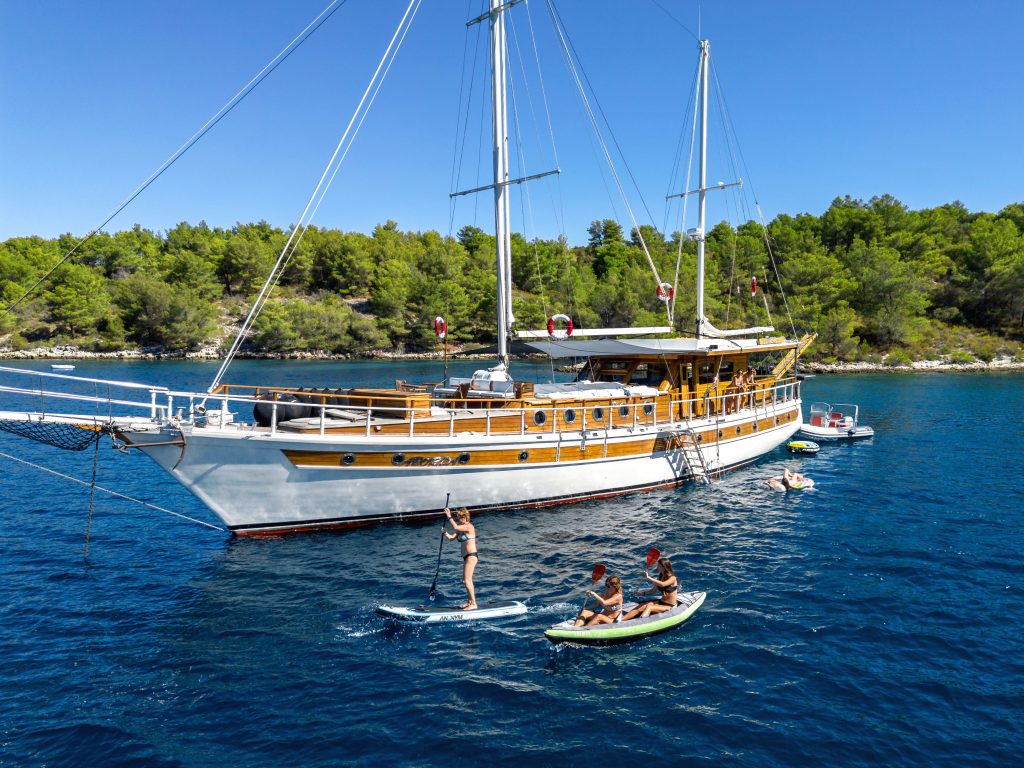 ABORDA A large wooden sailboat is anchored in clear blue water near a forested shoreline. People are paddleboarding and kayaking around the boat, enjoying the sunny day.