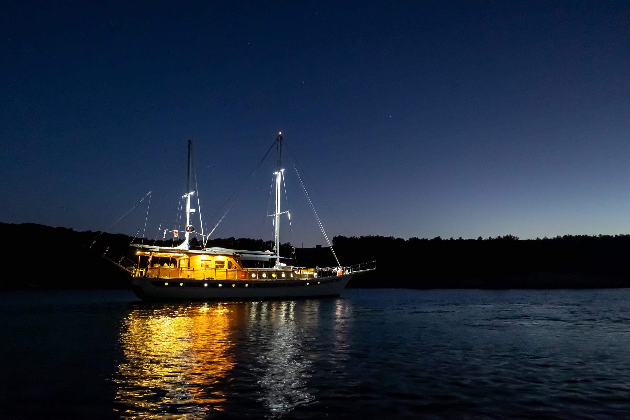 ABORDA A sailboat with its lights on is anchored on calm water at dusk, reflecting warm yellow light on the surface, with a dark shoreline and clear evening sky in the background.