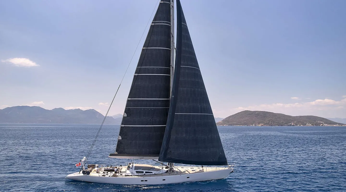 A sleek white sailboat with large black sails glides across calm blue water, with distant mountains and small islands visible—an ideal scene for a memorable yacht charter under a clear, sunny sky.
