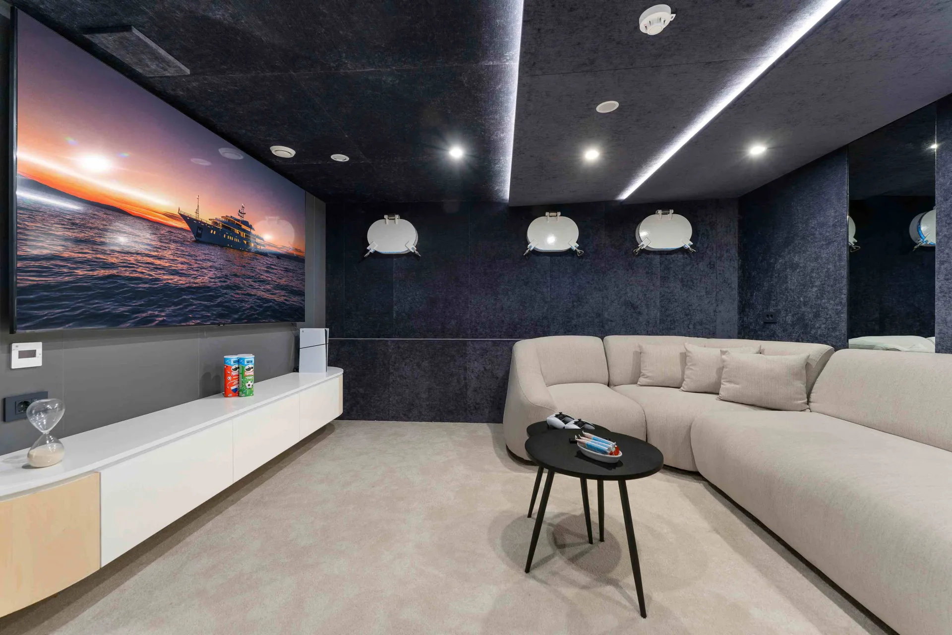 A modern home theater room with a large screen displaying a ship at sunset, a curved beige sofa, a small black table with remote controls, and shelves with snacks against dark walls and a light carpeted floor.