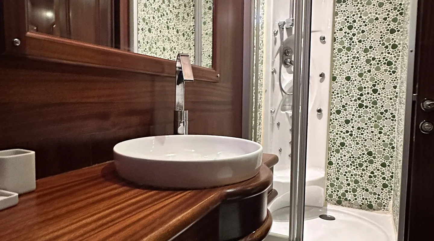 A modern bathroom with a round white vessel sink on a wooden countertop, a tall faucet, a glass shower enclosure, and mosaic tile walls in green and white tones. A mirror is above the sink.