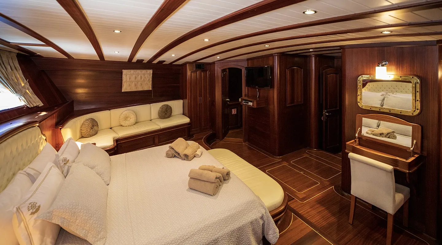 Luxurious yacht cabin with a large bed, cream bedding, rolled towels, sofa, vanity table with mirror, and elegant wooden walls and ceiling. Warm lighting creates a cozy and inviting atmosphere.