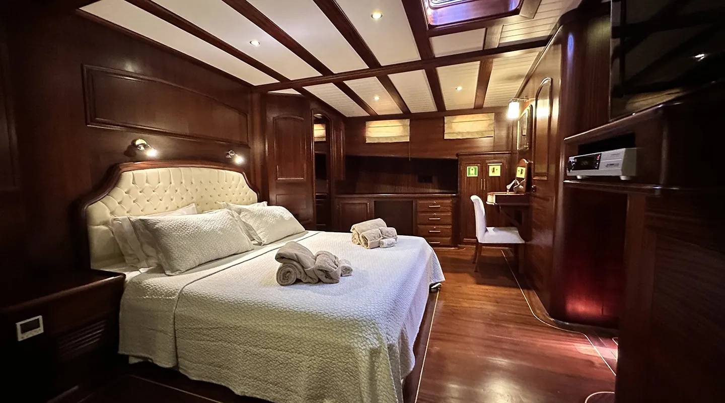 Luxurious bedroom with dark wood paneling, a large bed with white bedding and neatly folded towels, built-in cabinets, a desk with a chair, and warm ambient lighting.