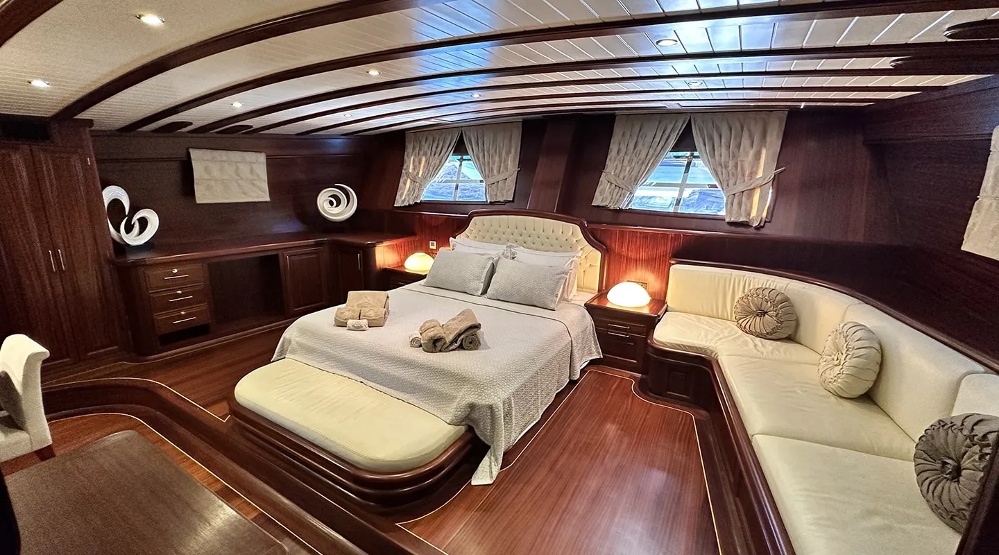 Luxurious yacht cabin with polished wood walls and ceiling, a large bed with light bedding, soft lighting, sculptural decor, two windows with drapes, and a cream-colored cushioned seating area wrapping around the corner.