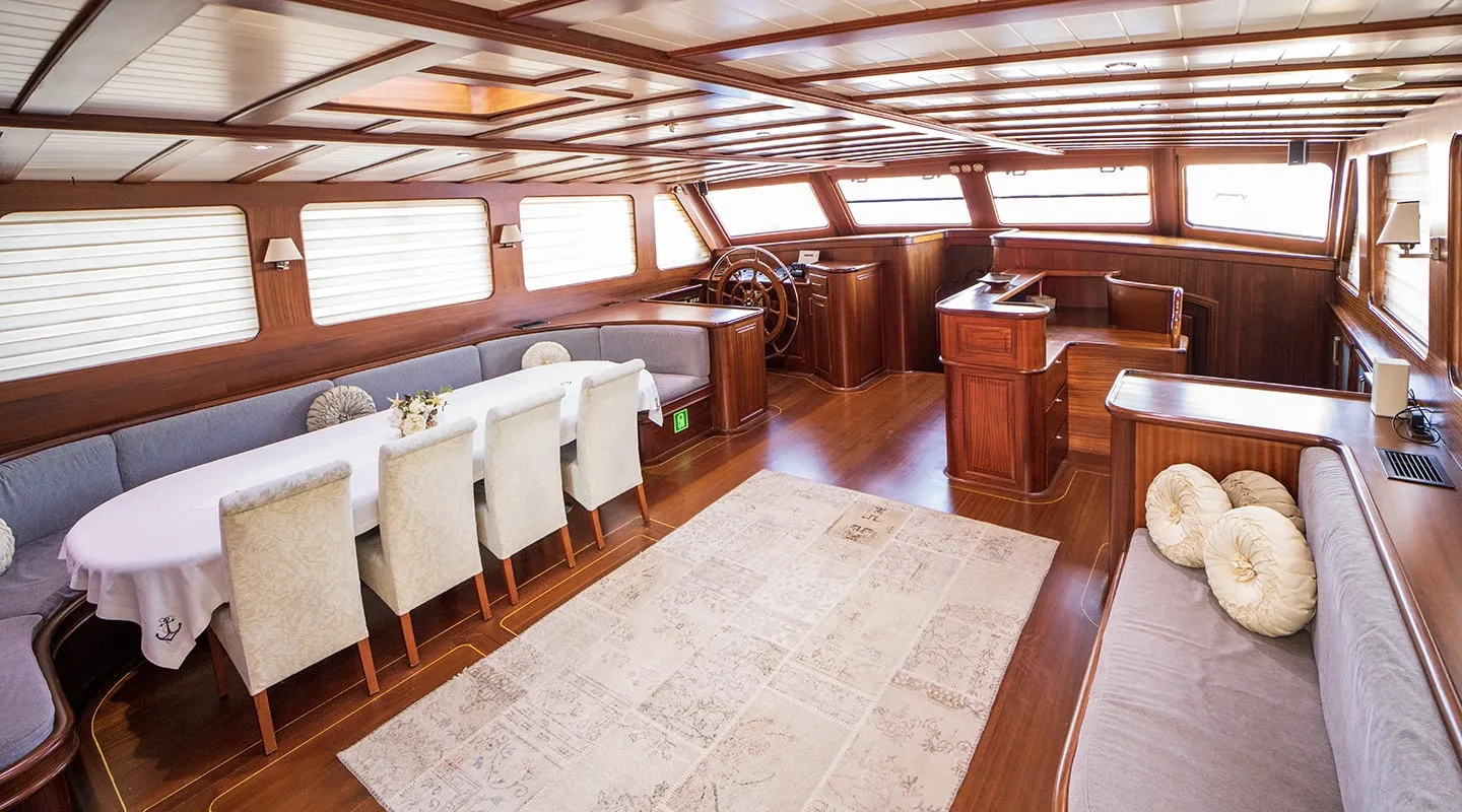 Spacious yacht interior with wooden floors and walls, featuring a round dining table with six chairs, a large steering wheel, cushioned benches, and large windows allowing natural light.