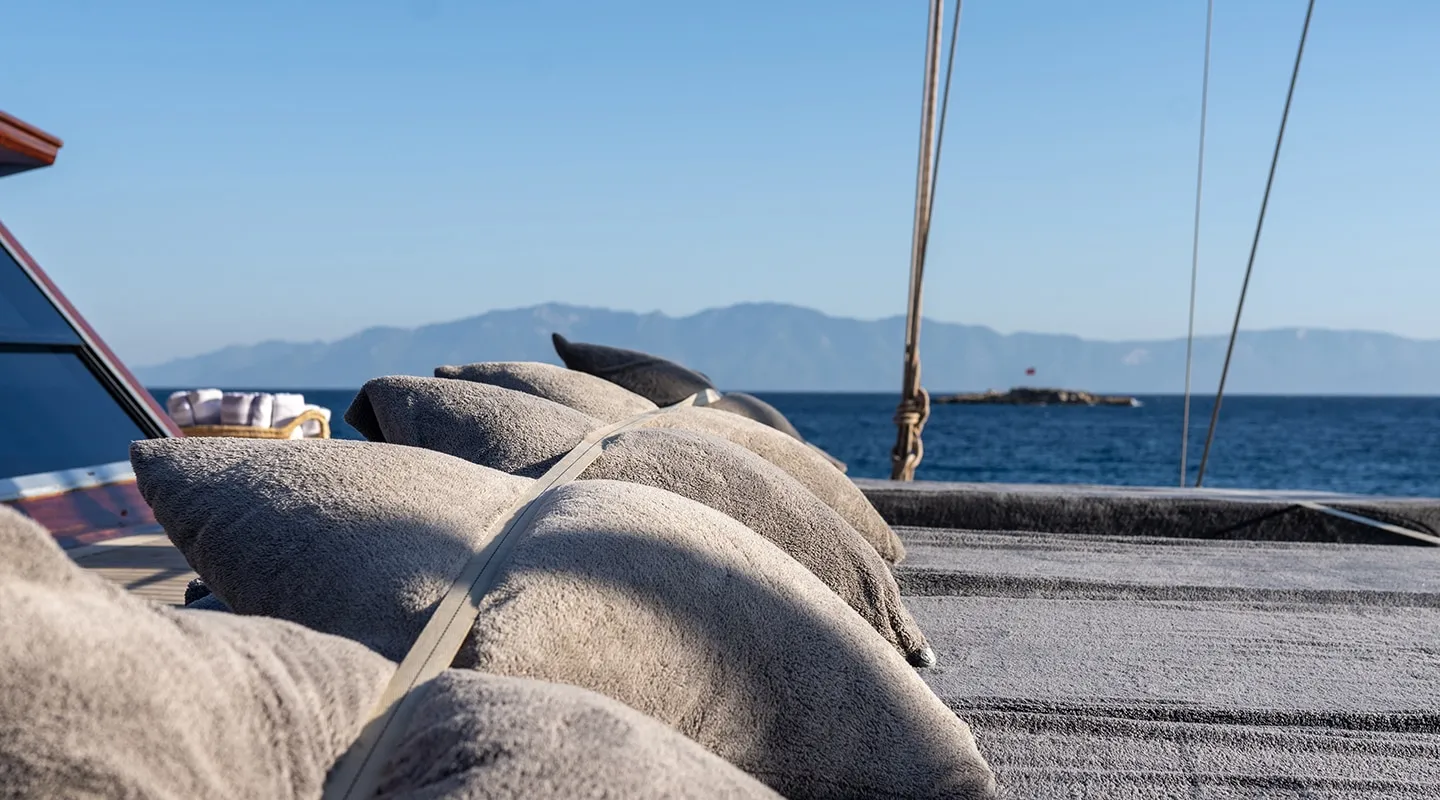 Several gray cushions are arranged on the deck of a boat with a strap securing them. In the background, calm blue sea and distant mountains are visible under a clear sky.
