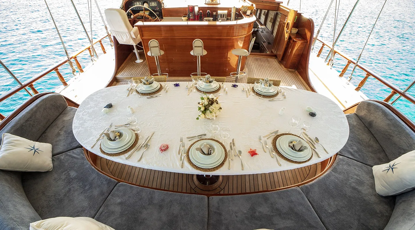 View of a yacht’s deck dining area with a table set for six, featuring elegant dishware, cutlery, and a floral centerpiece, surrounded by curved cushioned seating and overlooking blue water.