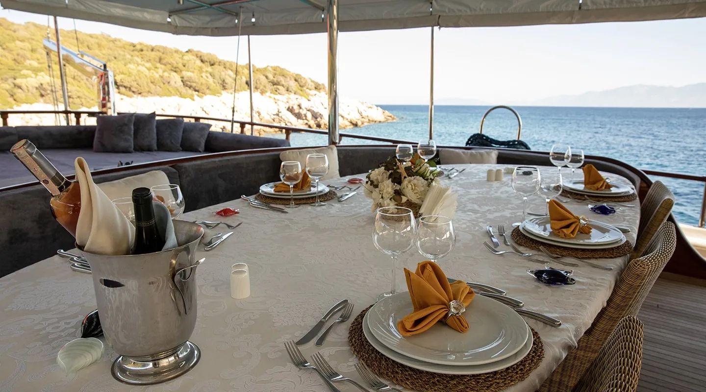 A table set for a meal on a yacht, with plates, glasses, silverware, and napkins, overlooks the sea and coastline. Bottles sit in an ice bucket and a floral centerpiece decorates the table.