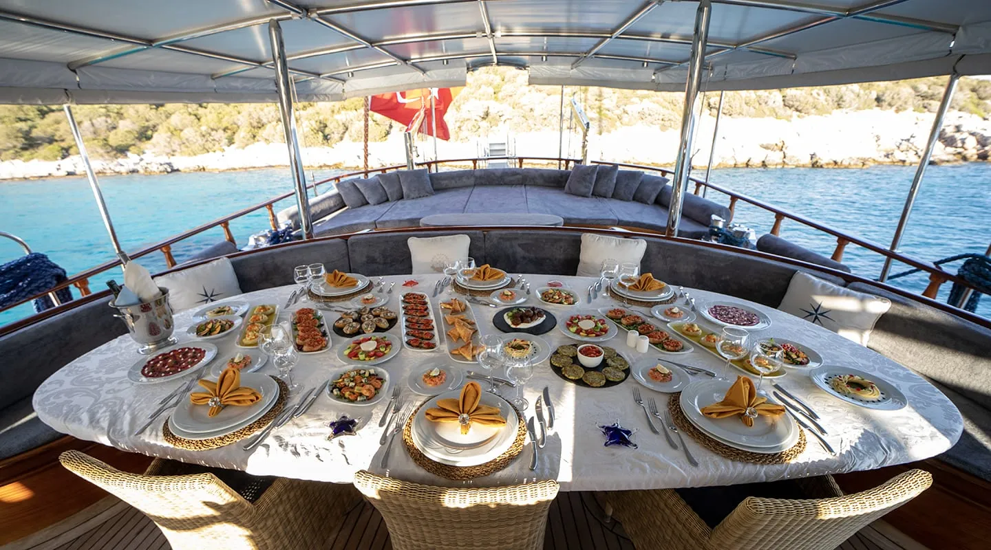 A luxurious yacht dining table is elegantly set with fine china, gold napkins, and a variety of appetizers and dishes. The table overlooks clear blue water and a rocky shoreline, with cushioned seating and shaded cover above.