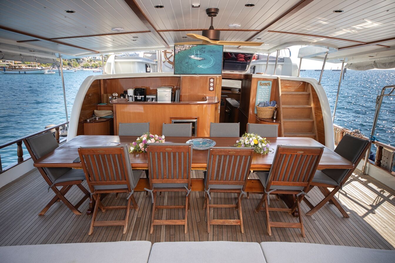 BODRUM QUEEN A wooden dining table with floral centerpieces and eight chairs is set on the deck of a yacht, overlooking the blue sea. The yacht’s interior includes a kitchenette, stairs, and a mounted TV.