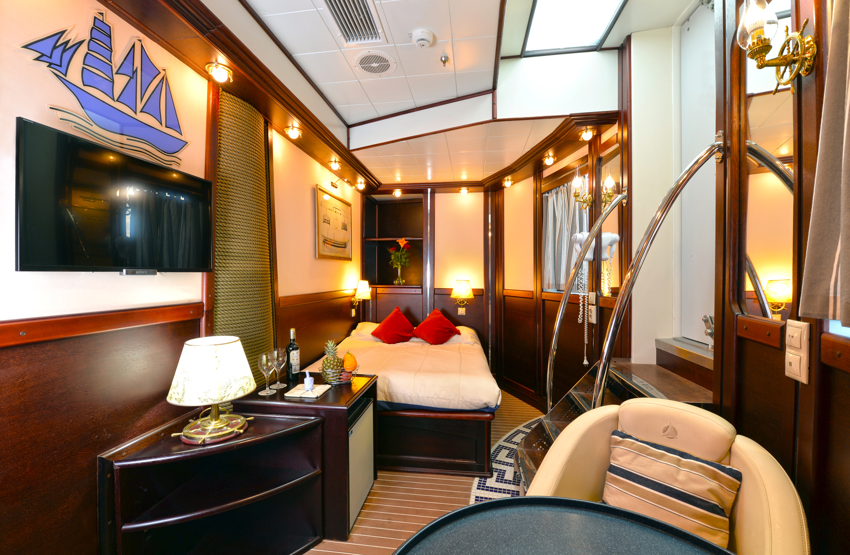 A cozy, nautical-themed hotel room with wood paneling, a double bed with red pillows, a flat-screen TV, a table with a lamp and wine, seating area, and a ship wall decoration. Warm lighting creates a welcoming atmosphere.