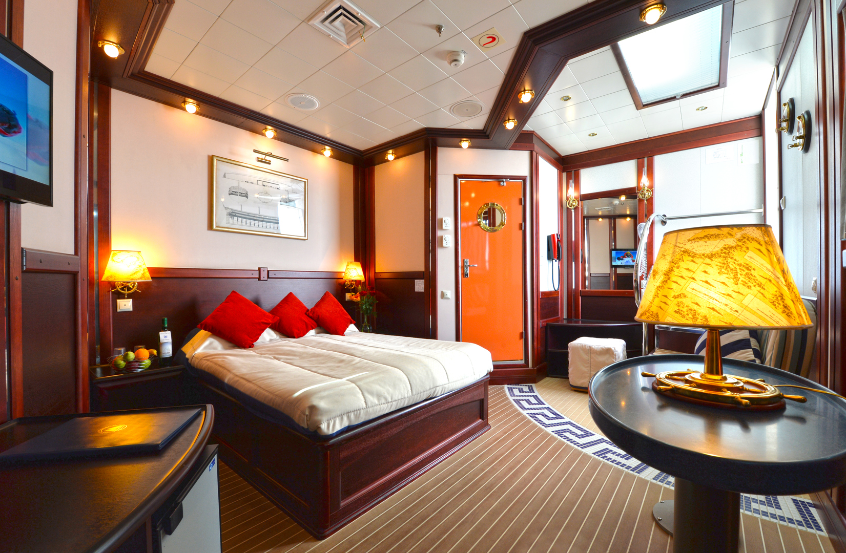 A cozy, well-lit cruise ship cabin with a double bed, red pillows, wooden furniture, a TV, a desk, a lamp with a map design, and a bright orange door. The room has nautical decor and modern amenities.
