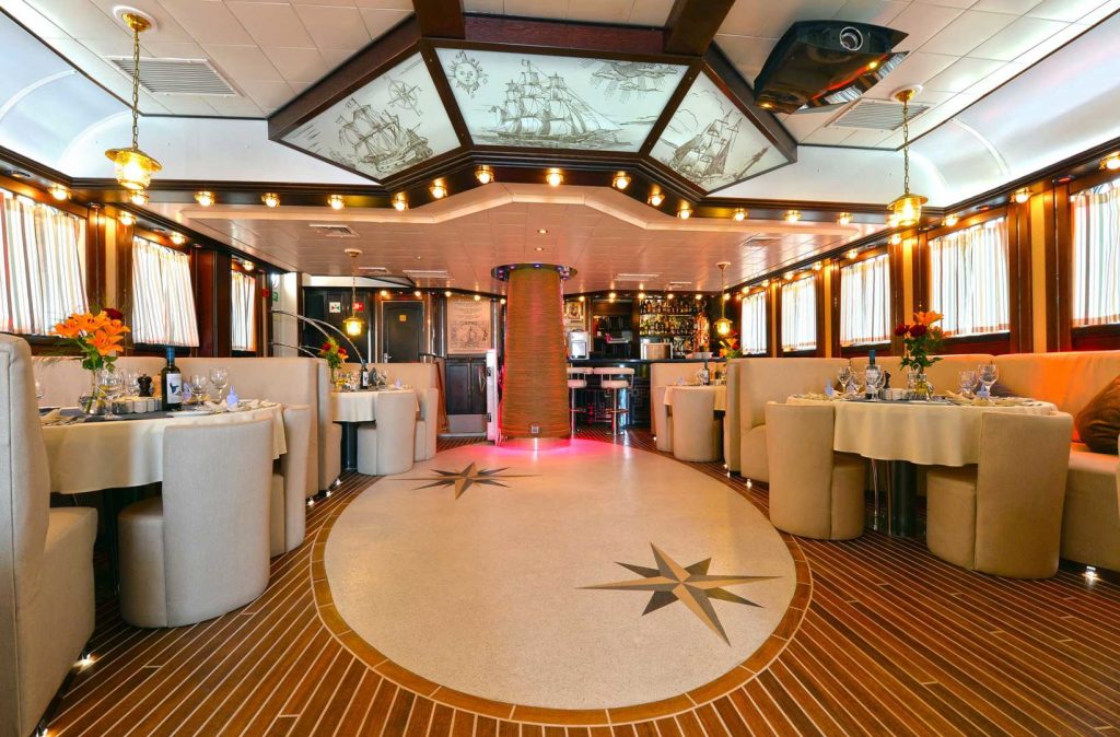 Elegant restaurant interior with cream-colored booths, round tables set with flowers and glassware, wood accents, nautical decor, and compass rose designs on the floor. Ceiling panels display ship illustrations. Warm, ambient lighting.