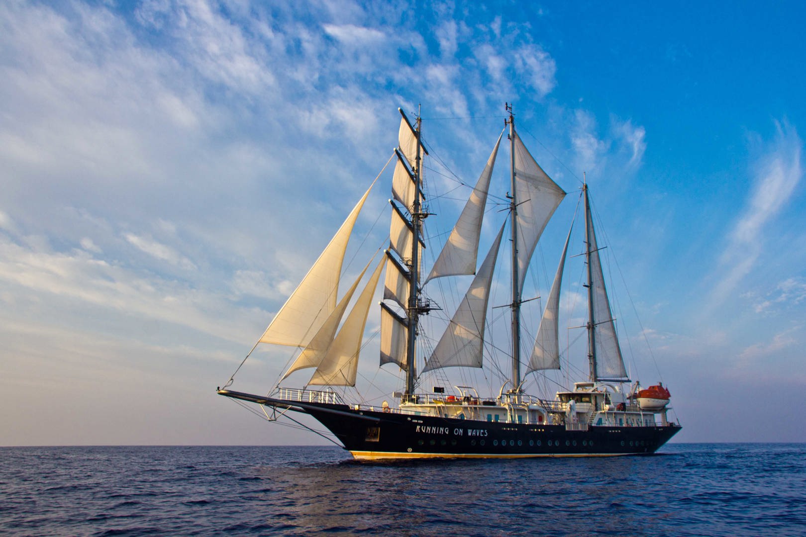 A large sailboat with white sails glides across calm blue water under a partly cloudy sky, with RUNNING ON WAVES written on its dark hull.