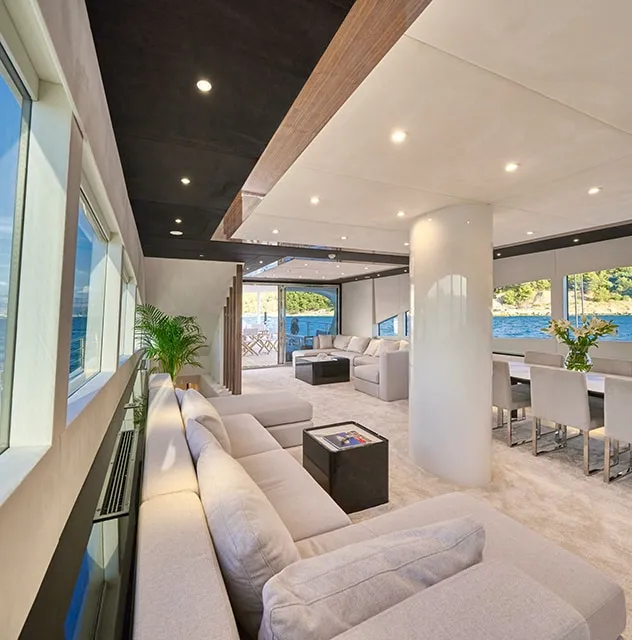 Luxurious yacht interior with large windows, white and beige sofas, a dining area, potted plants, and views of blue water and green hills—perfect for your next yacht charter. The space is bright and modern with recessed lighting.