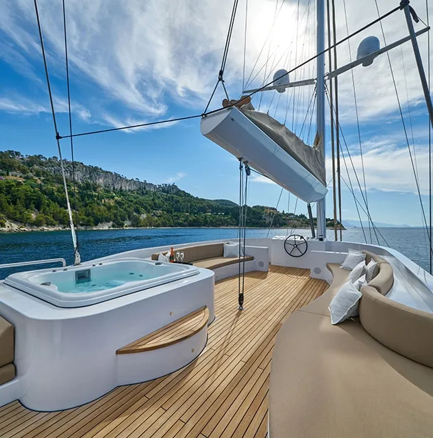 Luxury yacht deck with a hot tub, curved beige lounge seating, and scenic ocean views; ideal for a relaxing yacht charter with green hills and a rocky coastline visible under a blue sky with wispy clouds.