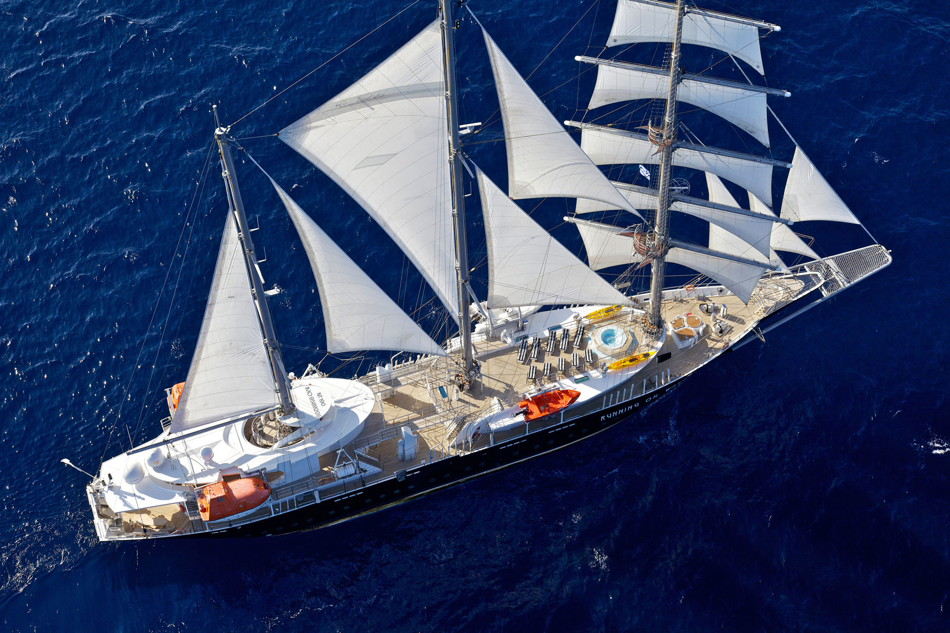 Aerial view of a large sailing yacht with multiple white sails open, cruising on deep blue water. The deck features sun loungers, a small pool, and lifeboats, with several people visible on board.