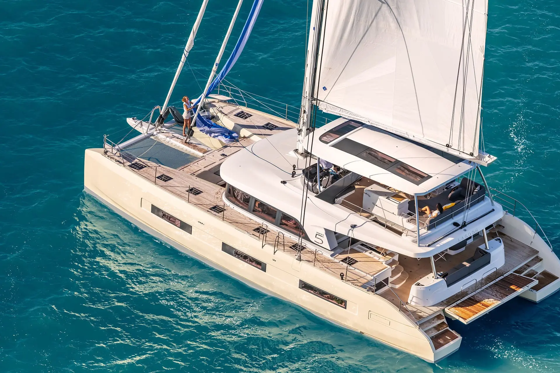 A white catamaran sailboat with people on board sails on clear blue water. The boat has multiple decks, a covered seating area, and open lounging spaces.