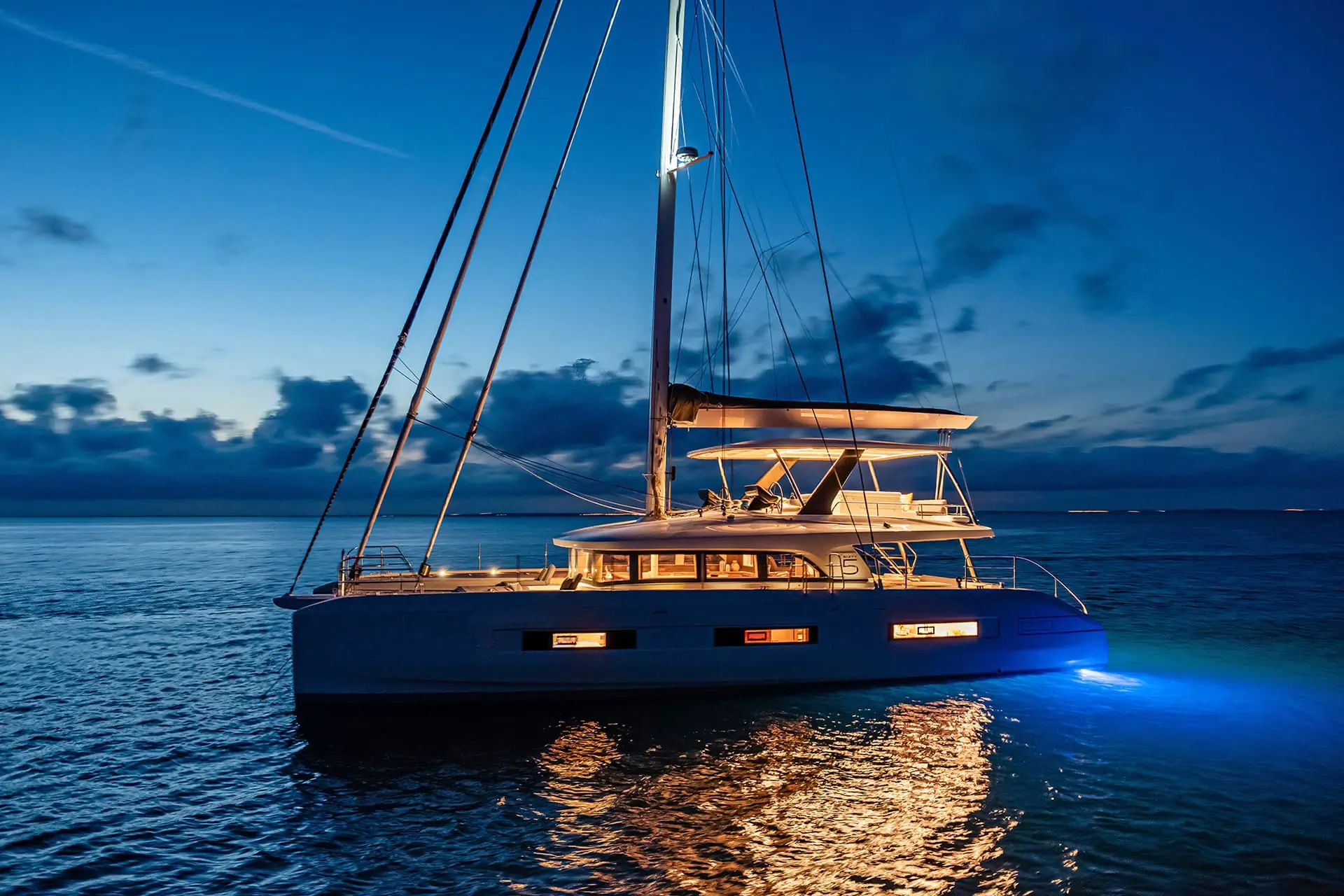 A luxury catamaran with illuminated windows and underwater lights floats on calm water at dusk, reflecting light on the surface under a dramatic evening sky.
