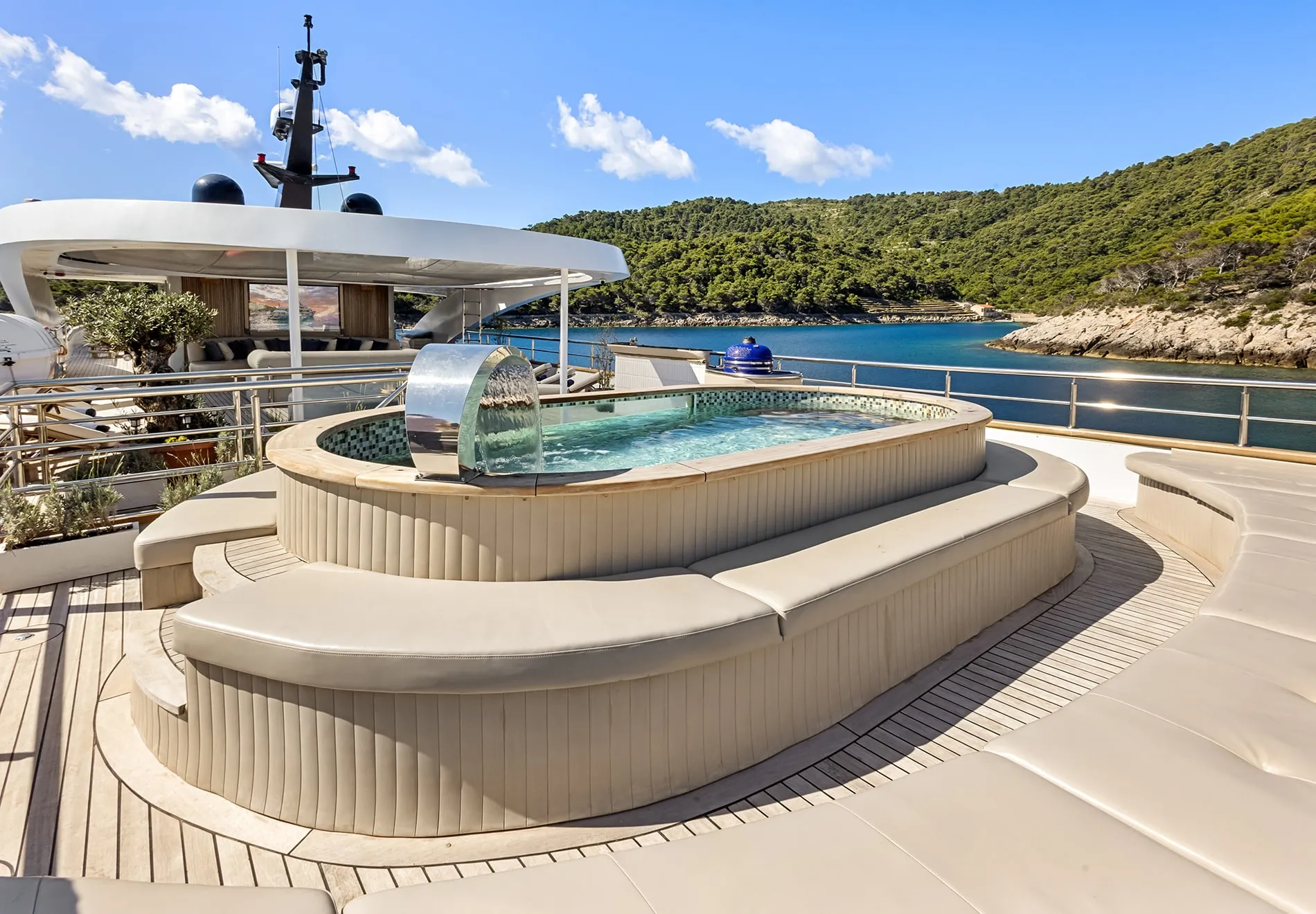A luxury yacht deck featuring a round jacuzzi with a water feature and surrounding cushioned seating, overlooking calm blue water and green, tree-covered hills under a clear sky.