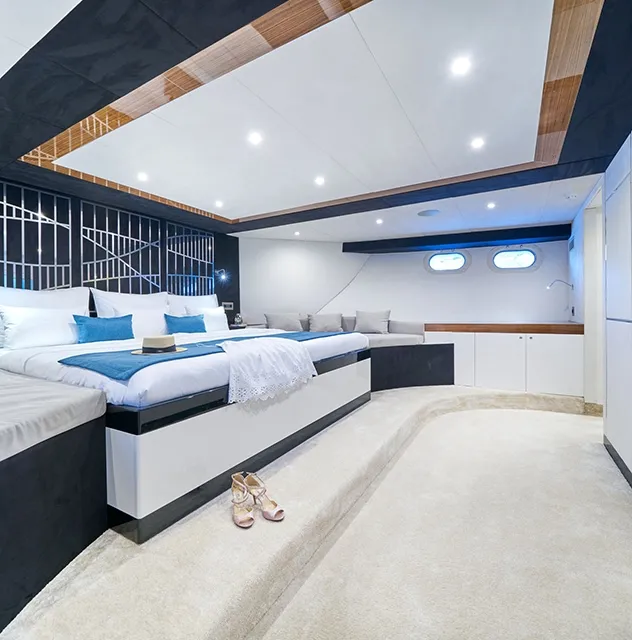 Spacious, modern yacht bedroom with a large bed, plush carpet, built-in sofa, decorative wall panels, and two round windows letting in natural light; perfect for relaxing during your luxury yacht charter. A pair of high-heeled shoes rests on the floor.