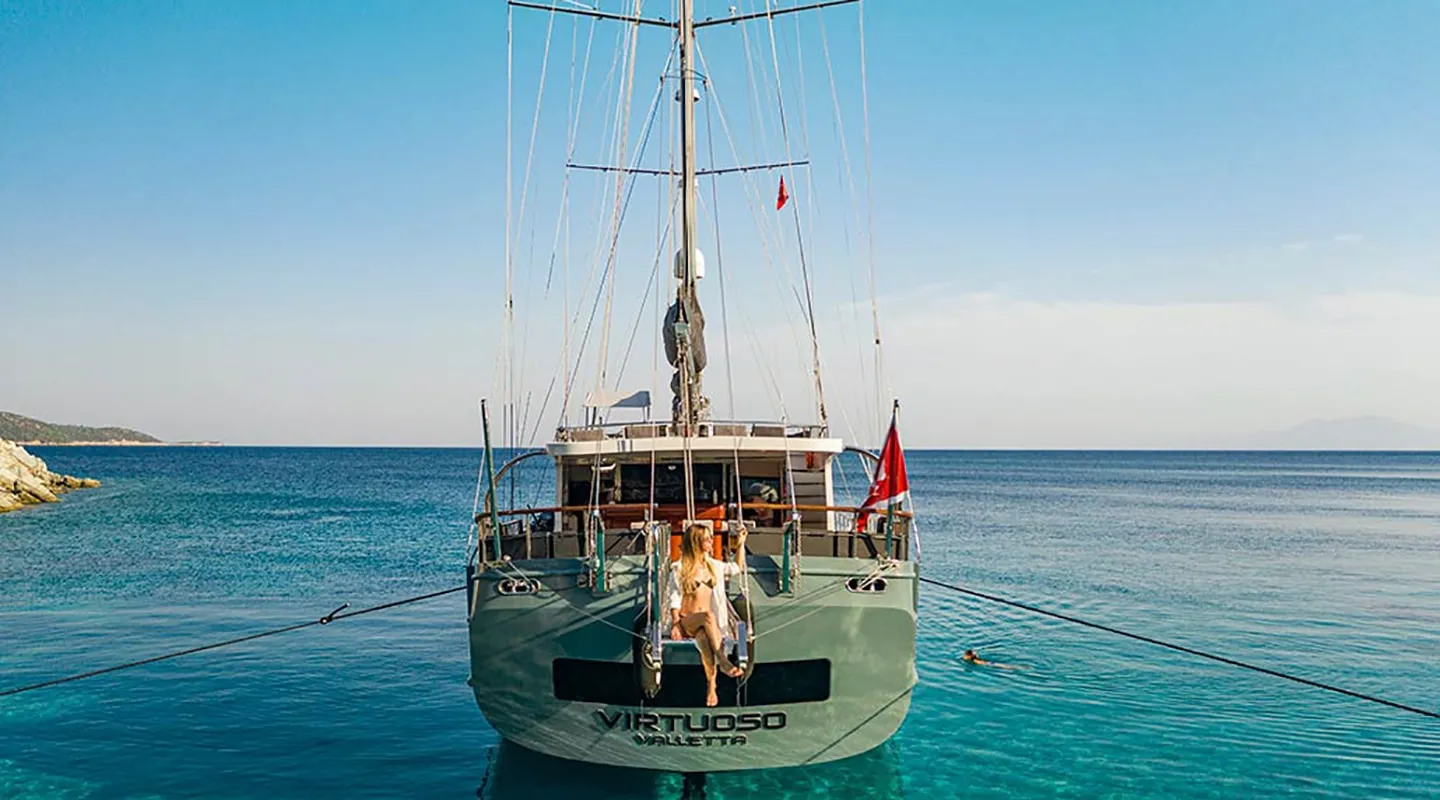 A person relaxes on the back deck of a moored sailboat named VIRTUOSO in clear, turquoise water, with a swimmer nearby and a rocky shore to the left under a clear blue sky.
