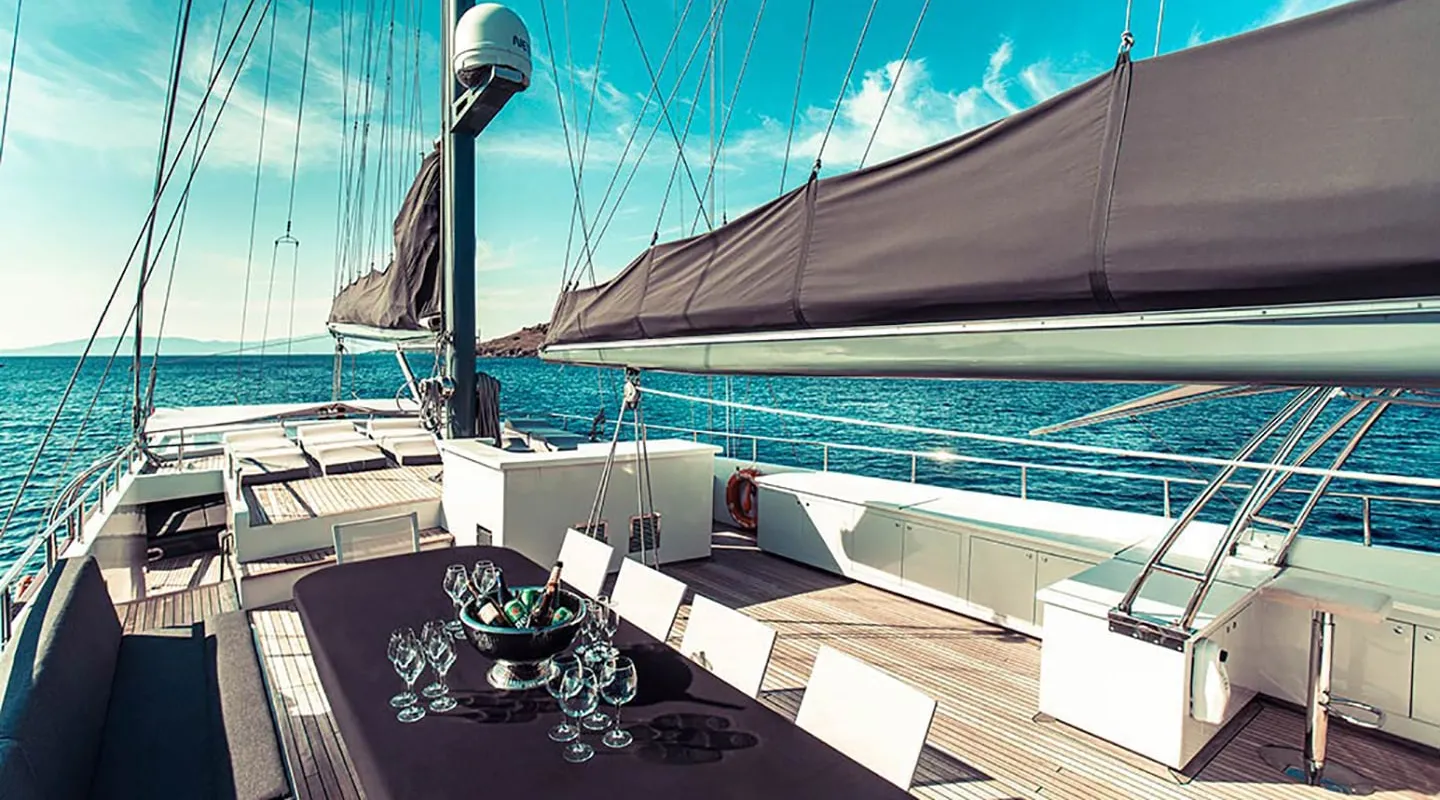 A luxury sailboat deck with a shaded dining table set with glassware, surrounded by white chairs, overlooking calm blue sea under a bright, clear sky.
