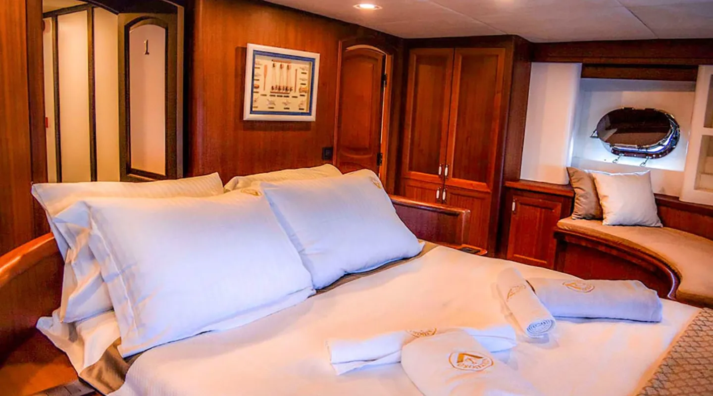 A cozy bedroom with wood-paneled walls, a large bed with white linens and pillows, folded towels on the bed, built-in cabinets, a small window seat with cushions, and a porthole letting in natural light.