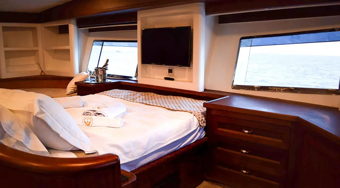A cozy yacht bedroom with a large bed, TV, wooden furniture, and two windows overlooking the ocean. Towels and a bottle of champagne on ice are placed on the neatly made bed.