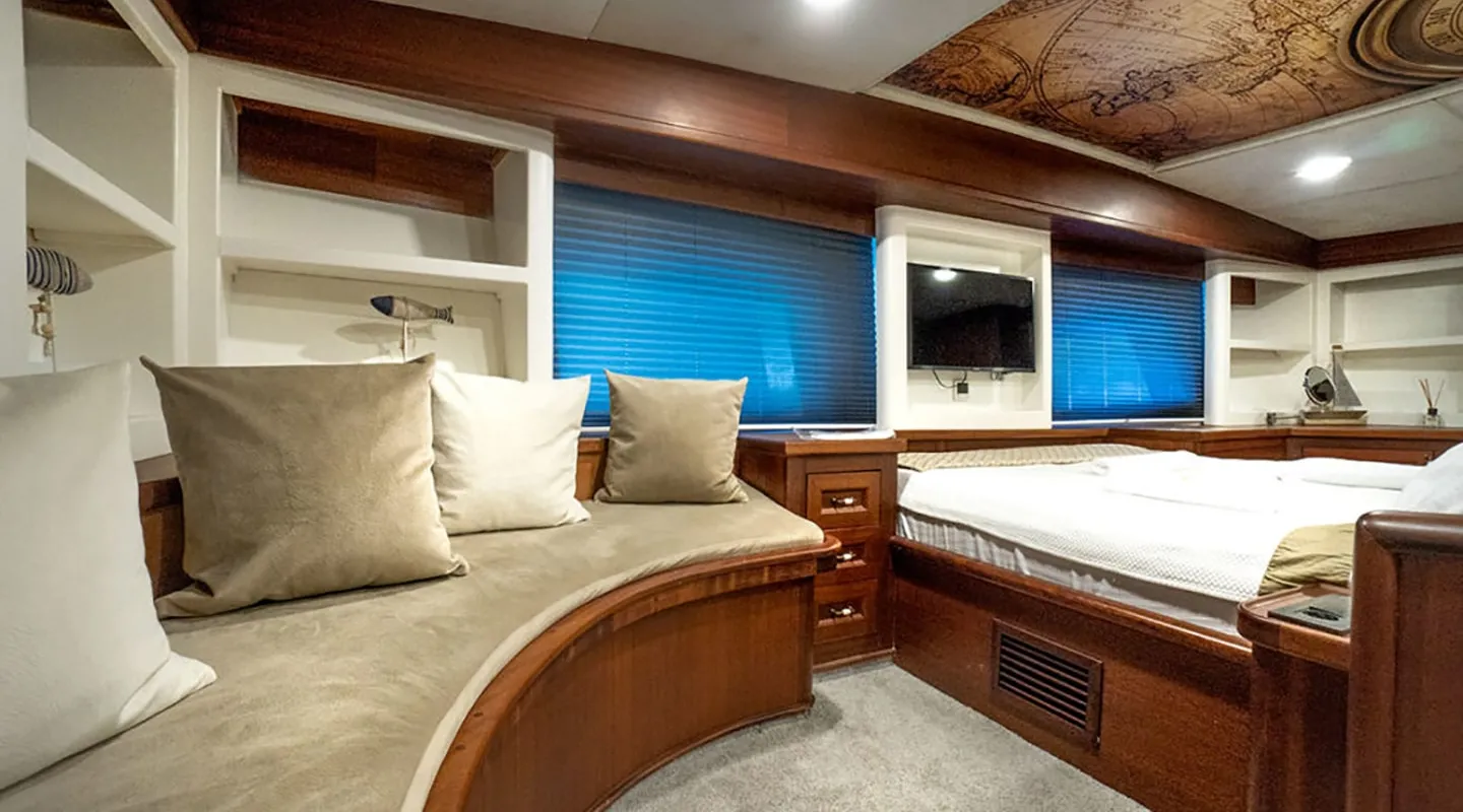 A cozy cabin interior features a curved wooden bench with beige cushions, a large bed, built-in white shelves, a wall-mounted TV, blue-tinted windows, and a ceiling with a vintage map design.