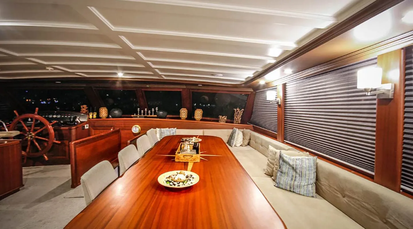 A luxurious yacht interior with a long wooden dining table, cushioned bench seating, chairs, decorative pillows, and nautical decorations, including a ship’s wheel and large windows with blinds.
