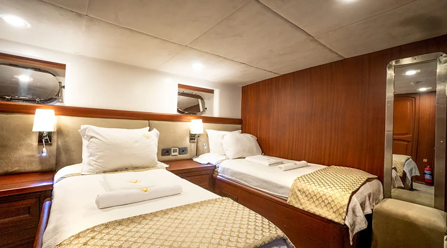 A cozy cabin with two single beds, each with white bedding and folded towels. Warm wood paneling, wall-mounted lamps, small windows, and a large mirror create an inviting, well-lit space.