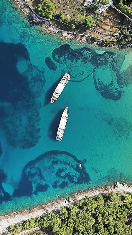 Aerial view of two boats anchored in clear turquoise water near a rocky, tree-lined shoreline with visible underwater reefs.