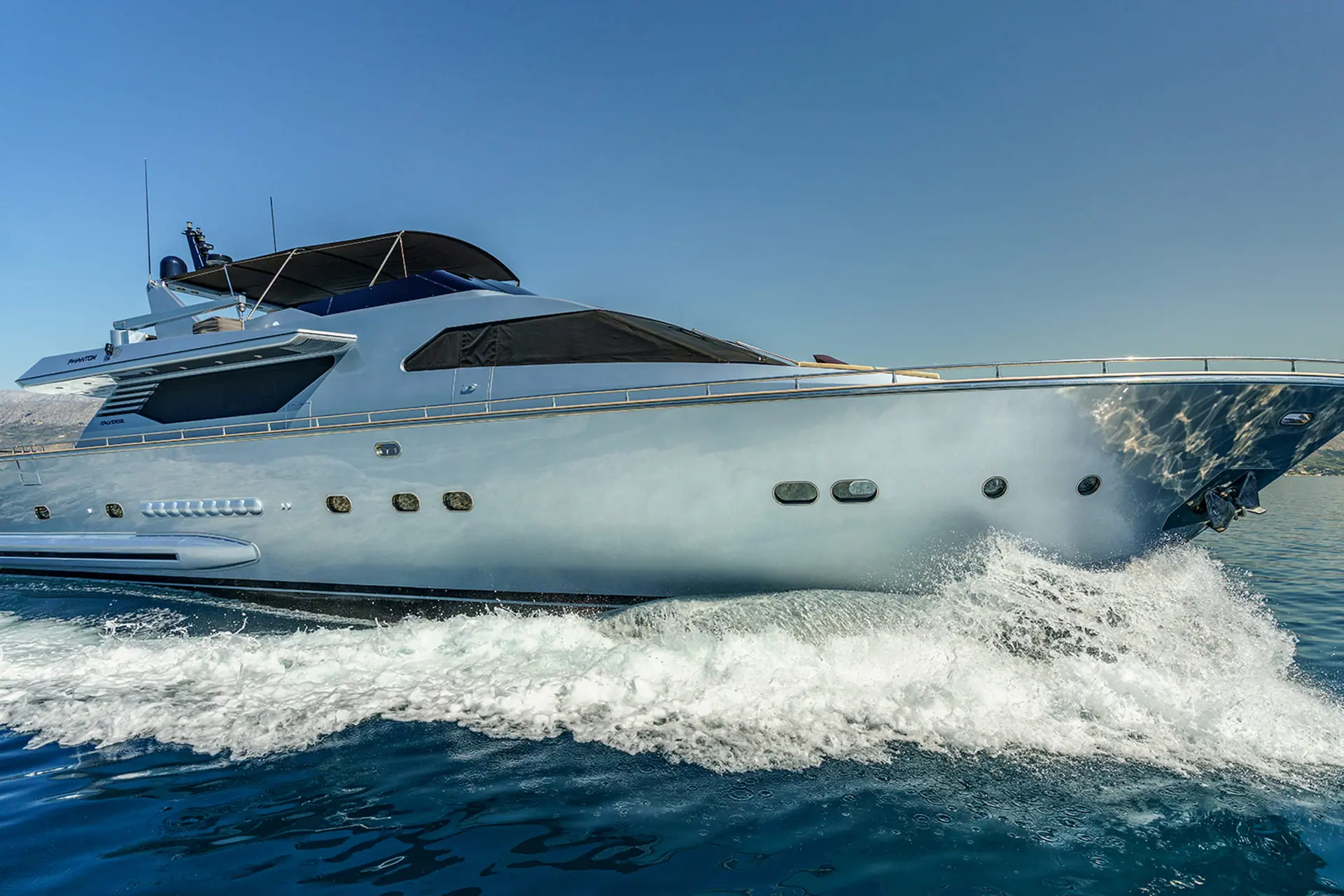 A sleek, modern yacht cruises quickly through calm blue water, creating white waves along its hull under a clear, sunny sky.