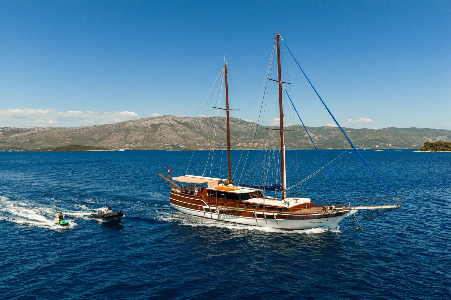 ELEGANZA A wooden sailboat with two masts sails on deep blue water near a mountainous coastline under a clear sky. A person rides a jet ski alongside the boat, creating a trail in the water.