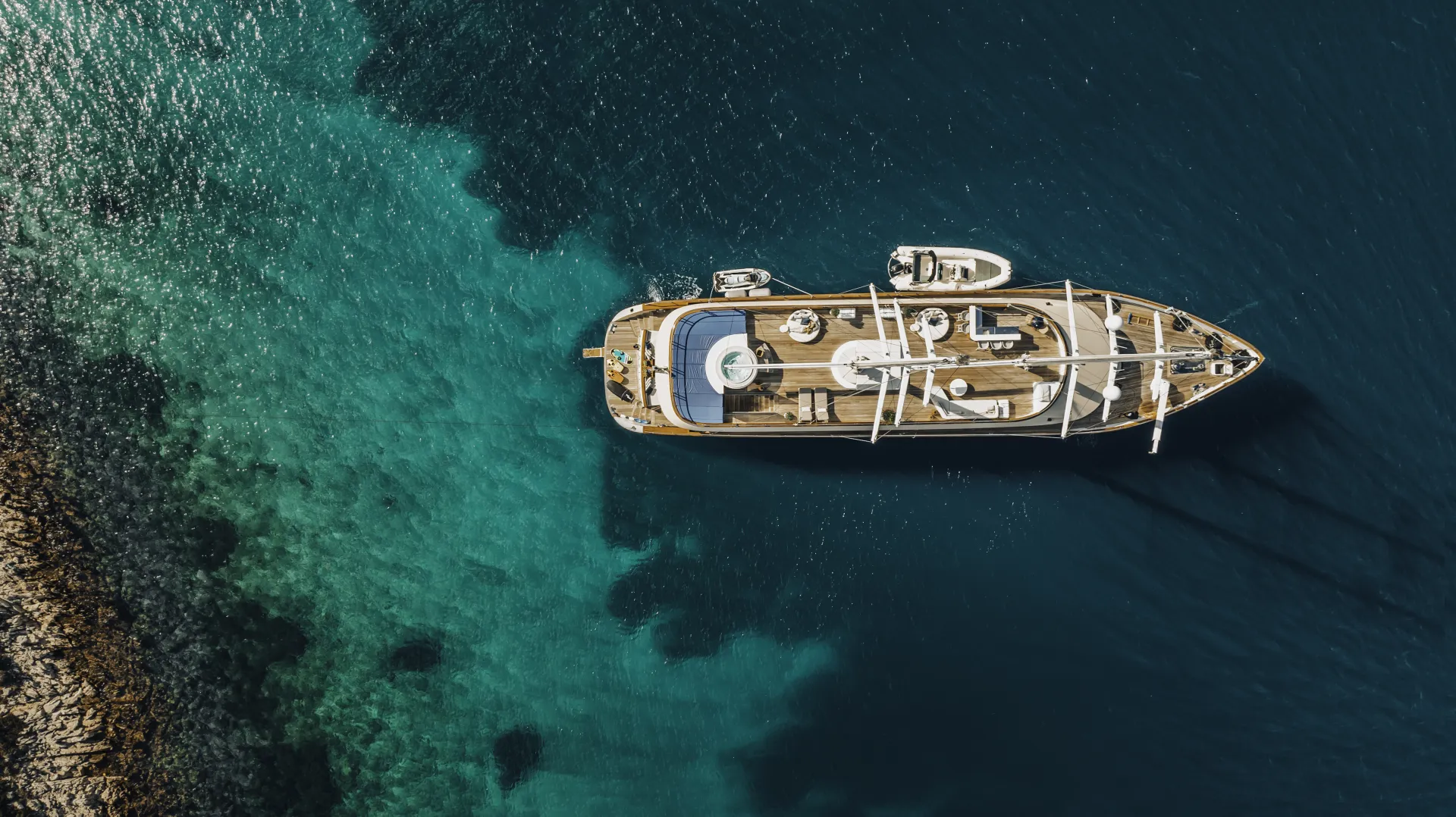 MAXITA Aerial view of a large yacht anchored in clear blue water near a rocky shoreline, with smaller boats visible on the deck and light and dark shades in the sea indicating varying depths.