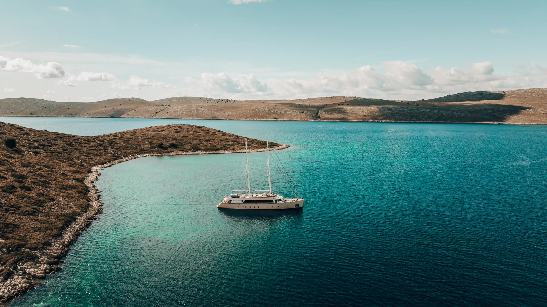 MAXITA A sailboat anchored in clear turquoise water near a small, hilly island under a blue sky with scattered clouds. Rocky shoreline and distant hills are visible in the background.