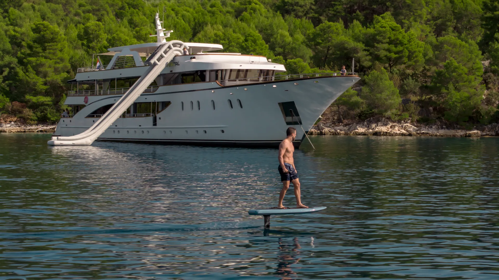 CRISTAL A man rides an electric hydrofoil surfboard on calm water near a large white yacht with a waterslide, anchored by a green, tree-lined shore.