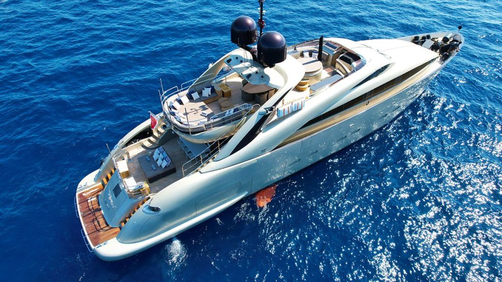 YCM 120 Aerial view of a sleek, modern luxury yacht with multiple decks floating on deep blue, clear ocean water under bright sunlight.