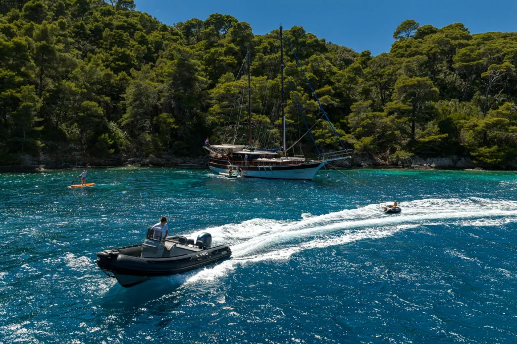 ELEGANZA A person drives a small inflatable motorboat on clear blue water near a forested shoreline, with a large sailing yacht anchored in the background and a paddleboarder nearby.
