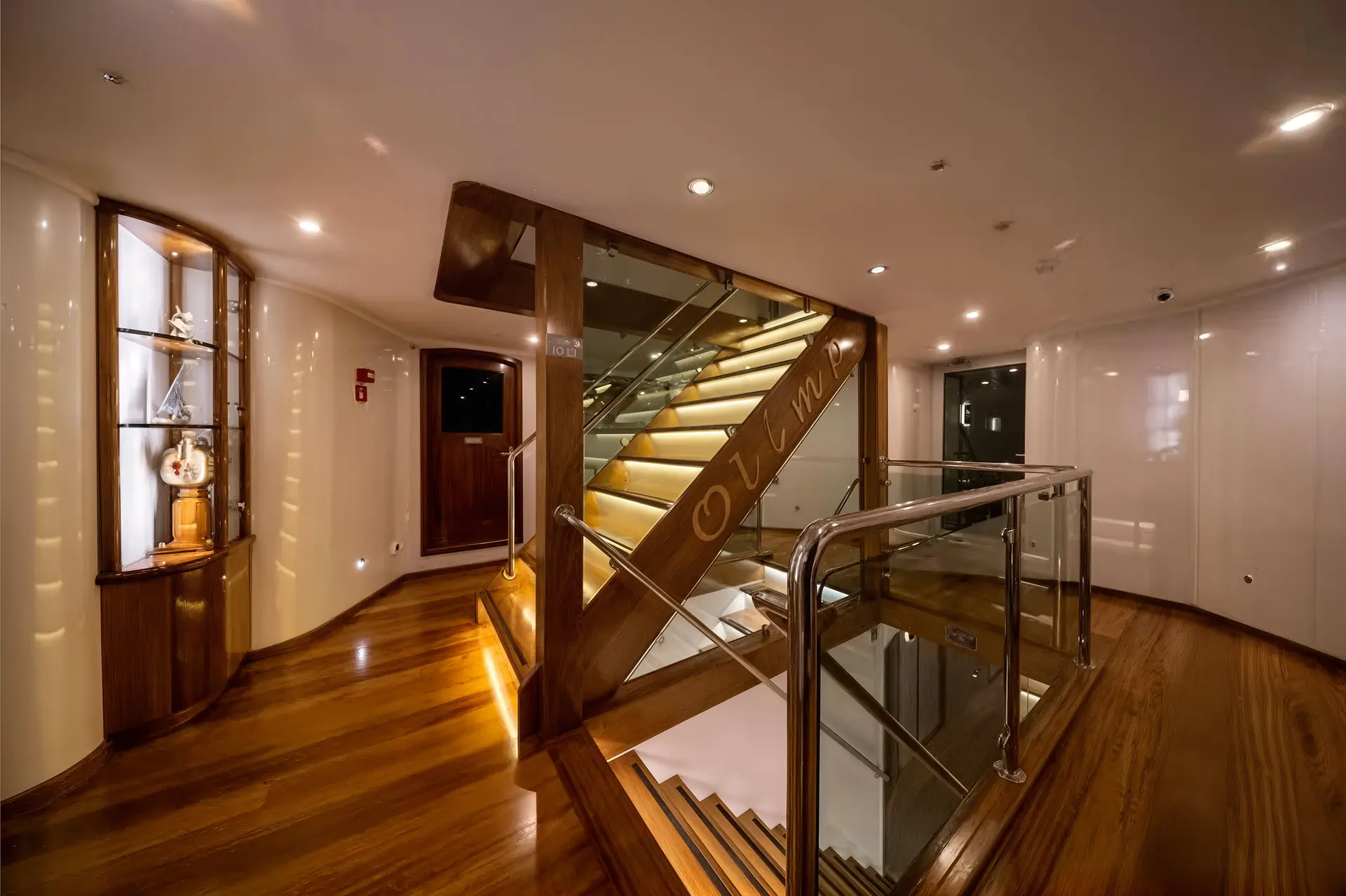 A luxurious yacht interior with polished wooden floors, a modern staircase with glass railings and illuminated steps, and display shelves holding decorative items.
