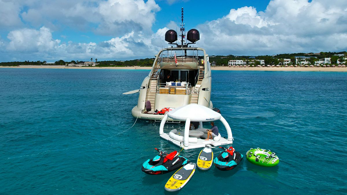 YCM 120 A luxury yacht is anchored in clear turquoise water near a coast. Several paddleboards, jet skis, and an inflatable lounge float beside the yacht. Houses and greenery are visible on the distant shoreline under a cloudy sky.