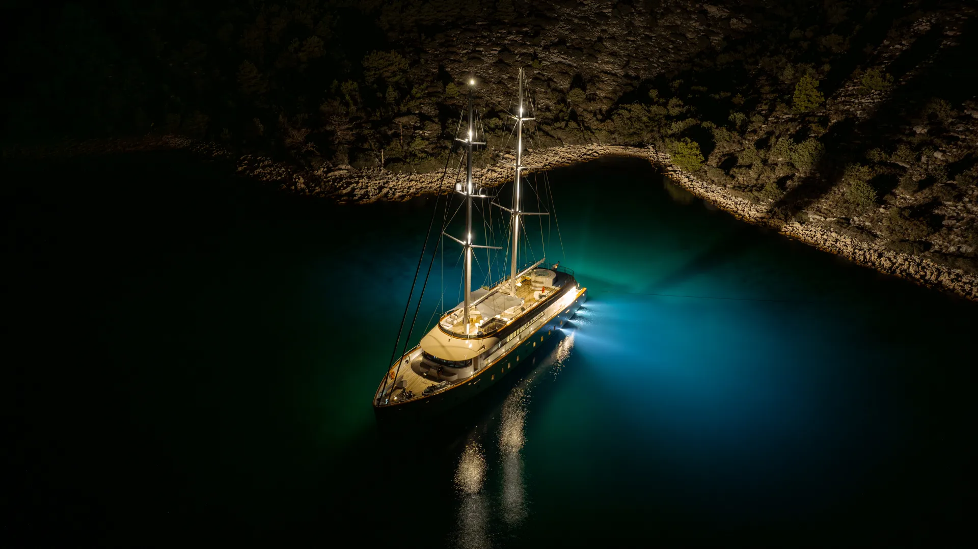 MAXITA A sailboat anchored in a calm, dark bay at night, illuminated by bright deck and underwater lights, with a rocky shoreline and trees visible in the background.