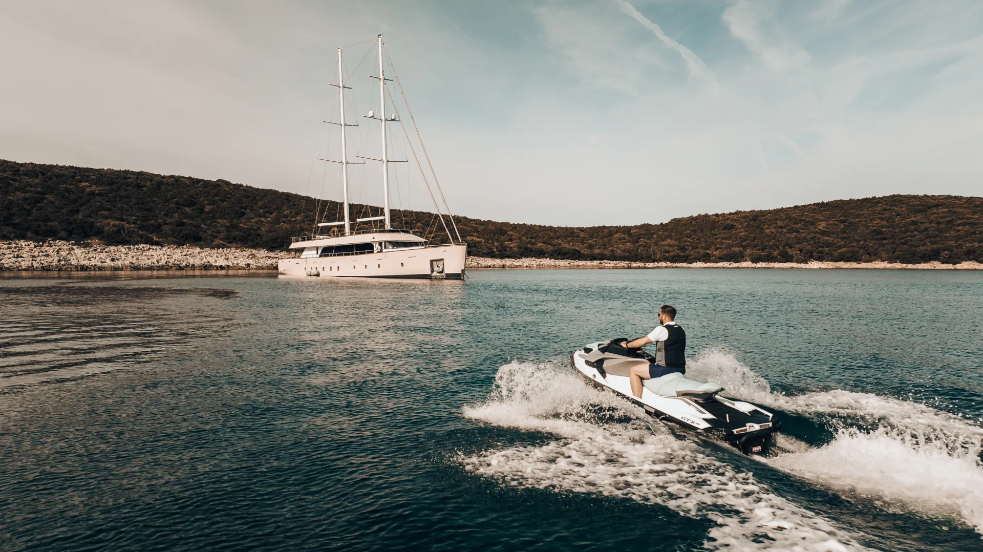 MAXITA A person rides a jet ski on calm blue water, heading toward a large white yacht anchored near a rocky shoreline with green hills under a partly cloudy sky.