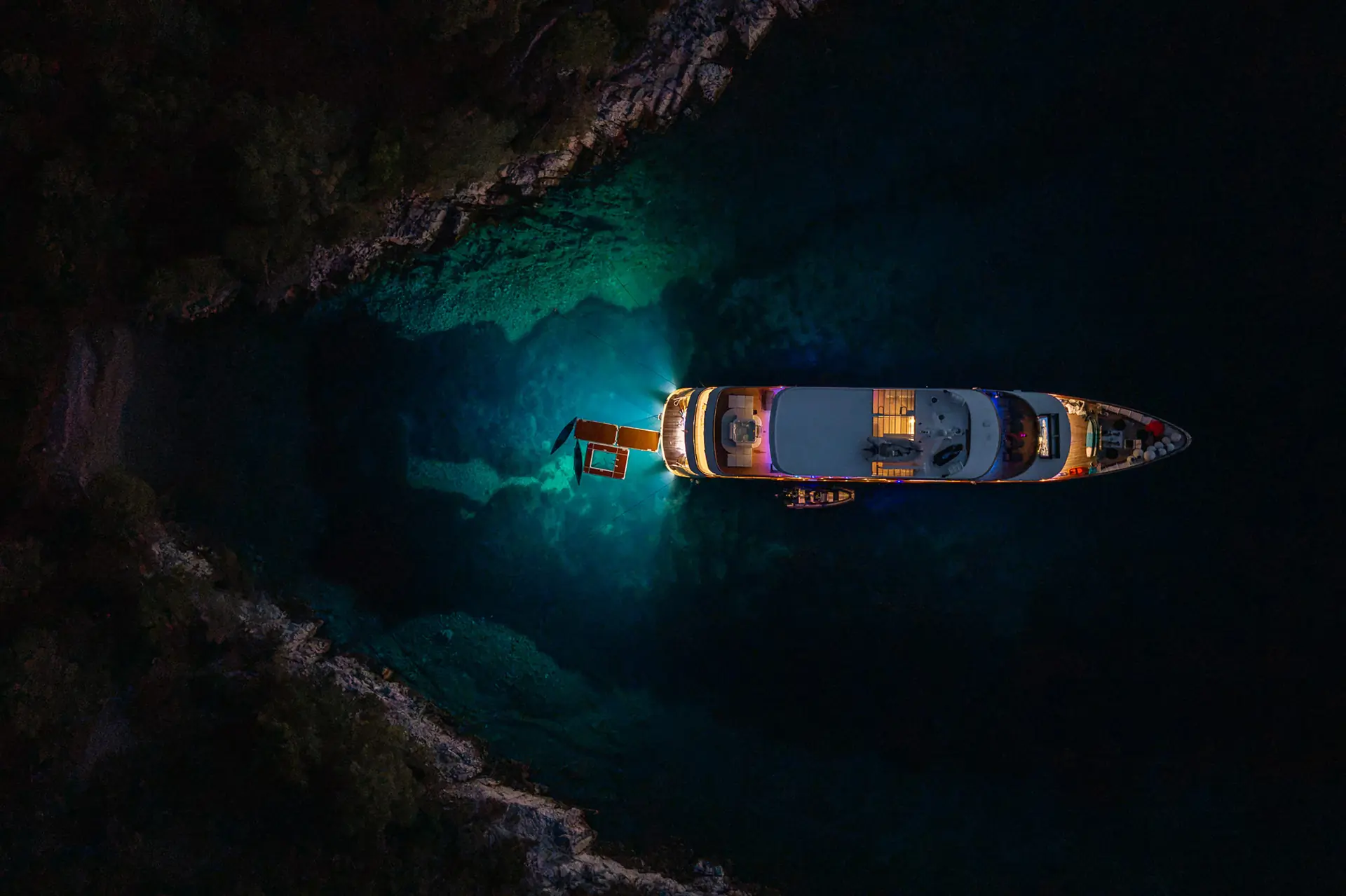 Aerial view of a lit yacht anchored in a narrow, rocky inlet at night, surrounded by dark water and illuminated turquoise water beneath the boat.