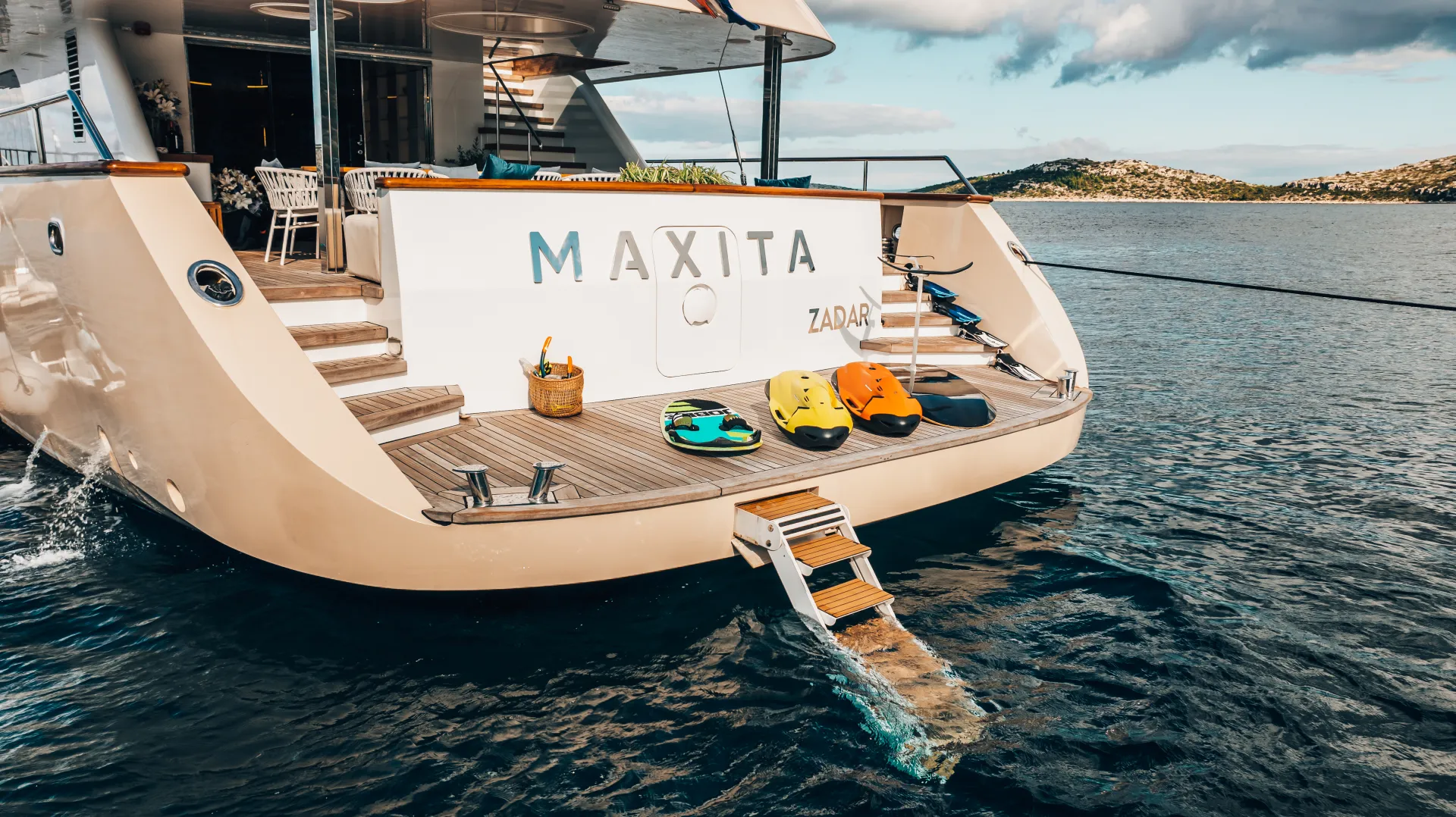 MAXITA The back of a luxury yacht named MAXITA with steps leading into the water, three colorful sea scooters, a basket with drinks, and a view of calm blue sea and distant islands.
