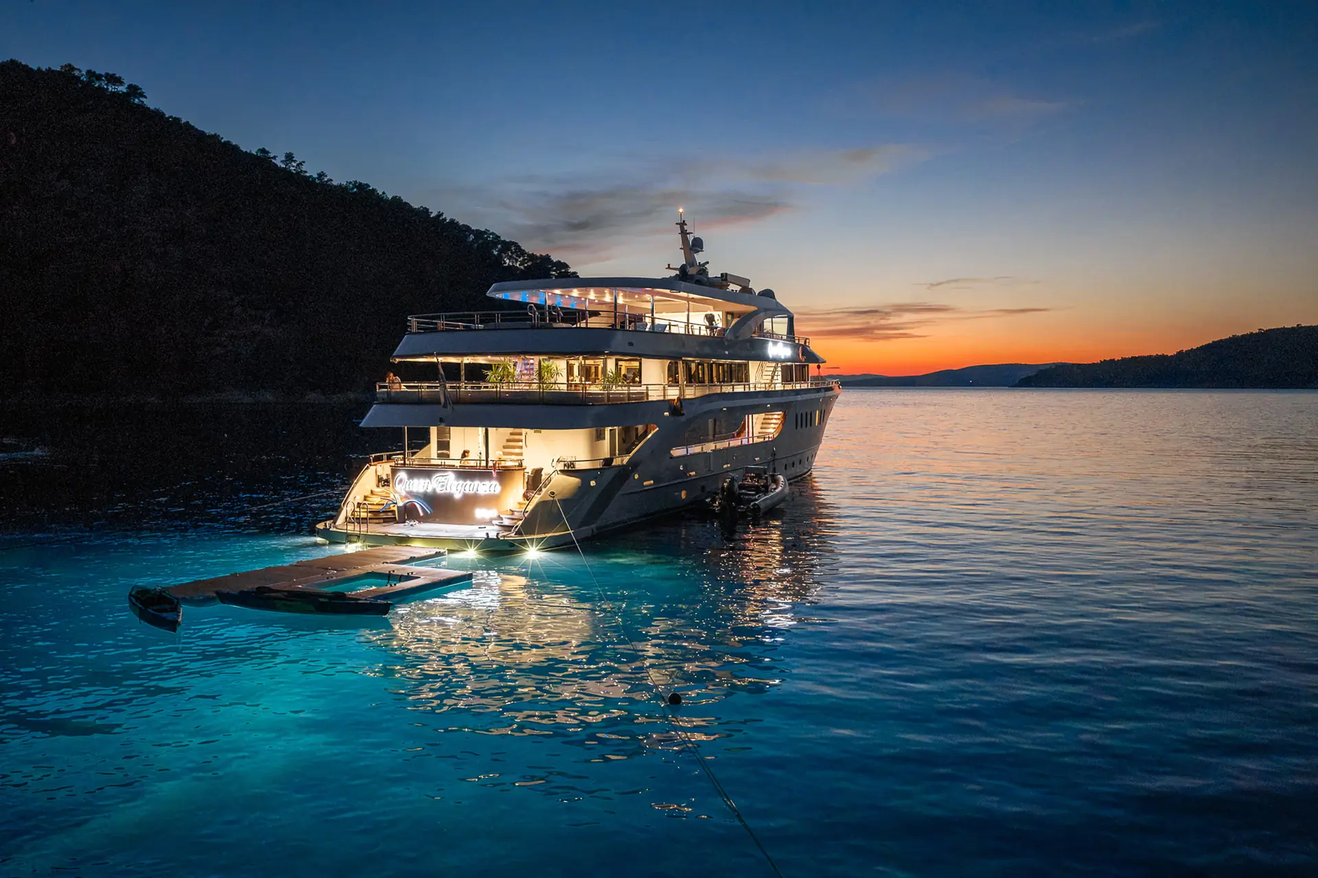 A luxury yacht is anchored in calm, blue water at sunset, with lights illuminating the stern and a floating platform nearby. Hills silhouette the horizon under a clear sky with orange and blue hues.